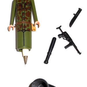 May include: A green plastic toy soldier with a camouflage pattern, holding a pen. The soldier is wearing a helmet and has a black face. The soldier is surrounded by black plastic toy weapons, including a knife, a rifle, and a baton.