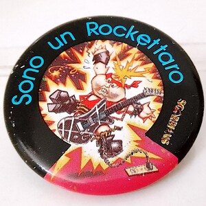 Puede incluir: Un botón redondo negro y rojo con un personaje de dibujos animados tocando una guitarra. El texto "Sono un Rockettaro" está escrito en azul alrededor del borde del botón. El texto "Snapper Bros" está escrito en blanco en el lado derecho del botón.
