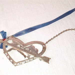 Op de afbeelding: Zilveren hartvormige ornament met een uitgesneden ontwerp van een stel dat elkaars hand vasthoudt en een pijl door het hart. De ornament heeft een blauw lint en een zilveren ketting.