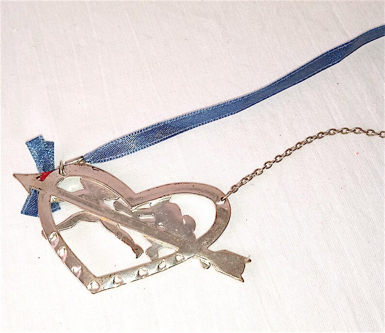 Op de afbeelding: Zilveren hartvormige ornament met een uitgesneden ontwerp van een stel dat elkaars hand vasthoudt en een pijl door het hart. De ornament heeft een blauw lint en een zilveren ketting.