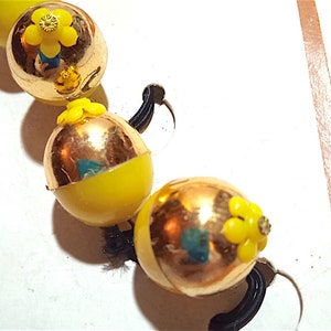 Puede incluir: Primer plano de un collar con cuentas amarillas y doradas. Cada cuenta tiene un adorno de flor amarilla. Las cuentas están ensartadas con un cordón elástico negro. El collar está sobre una superficie blanca.