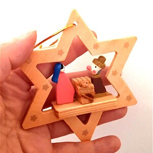 Può includere: Un ornamento di legno a forma di stella con un ritaglio della scena della natività. L'ornamento presenta una donna dai capelli castani con un vestito rosso, un uomo con una tunica e un cappello marroni, e una mangiatoia di legno con un bambino dentro. L'ornamento è decorato con stelle dorate.