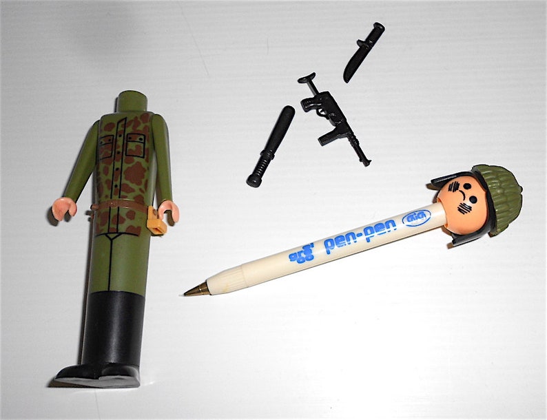 May include: A green camouflage military figure with a black belt and a brown pouch. The figure is missing its head. There is a black pen with a blue and white label that says "pen-pen" and a soldier head eraser on the end. There is also a black knife, a black baton, and a black rifle.
