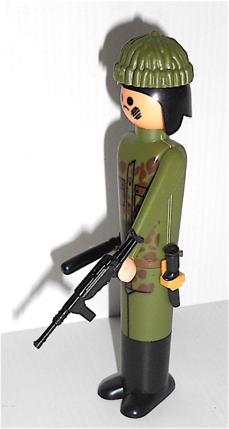 May include: A green camouflage plastic toy soldier with a black machine gun and a green knit hat. The soldier is wearing black boots.