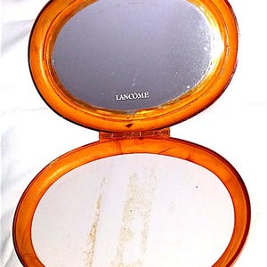 Può includere: Uno specchio compatto in plastica arancione con un interno bianco e la parola "LANCOME" stampata sullo specchio.