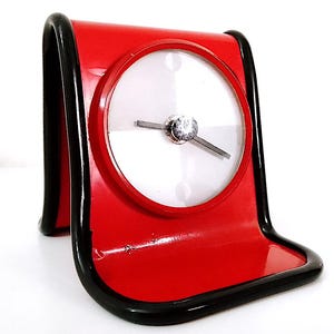 May include: A red and black desk clock with a white face and black hands. The clock is shaped like a wave and has a black base.