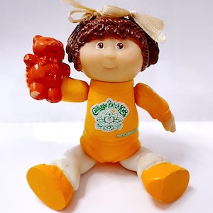 May include: A vintage Cabbage Patch Kids doll with brown curly hair and a cream-colored bow. The doll wears an orange outfit and holds an orange teddy bear. The doll's outfit has the Cabbage Patch Kids logo.