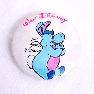 Könnte beinhalten: Ein weißer Knopf mit einer blauen und rosa Comicfigur mit Flügeln und Hasenohren. Die Figur lächelt und hat eine rosa Nase. Der Text "WALT DISNEY" ist in rosa um die Figur herum gedruckt.