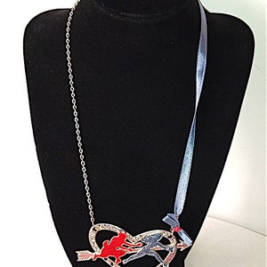 Op de afbeelding: Een zilveren ketting met een hartvormige hanger met twee figuren, een rode en een blauwe, die elkaar vasthouden en door de lucht vliegen. Het hart wordt doorboord door een pijl. De ketting heeft ook een blauw lint met een rode strik.