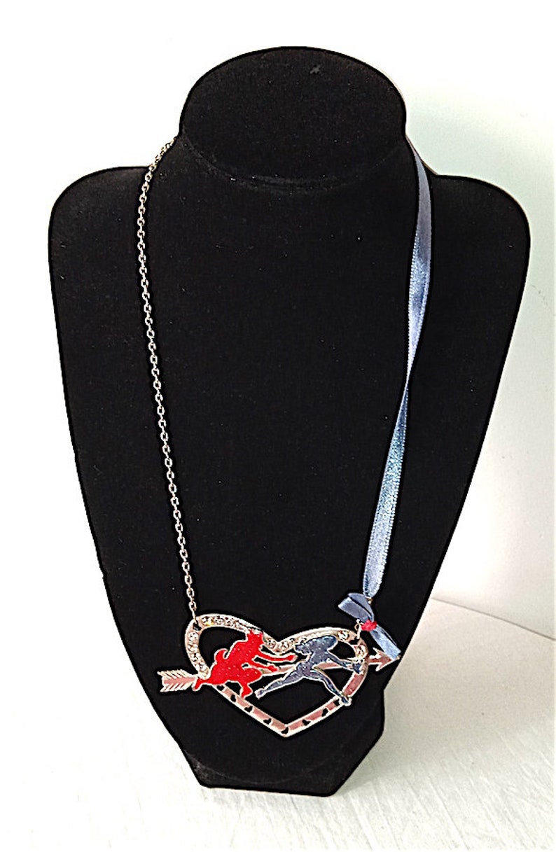 Op de afbeelding: Een zilveren ketting met een hartvormige hanger met twee figuren, een rode en een blauwe, die elkaar vasthouden en door de lucht vliegen. Het hart wordt doorboord door een pijl. De ketting heeft ook een blauw lint met een rode strik.