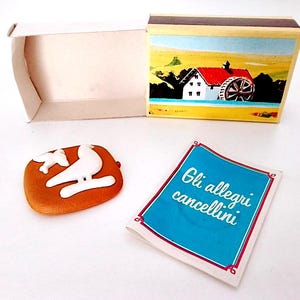 May include: A vintage matchbox containing a white bird eraser. The matchbox features a colourful illustration of a windmill and a house on the front. The eraser is shaped like a bird with a white body and brown wings. The matchbox also includes a small blue card with the text "Gli allegri cancellini" in white.