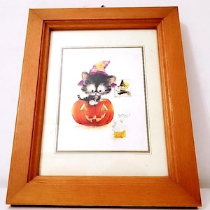 Può includere: Una stampa incorniciata di Halloween con un gatto nero che indossa un cappello da strega seduto all'interno di una zucca intagliata. Il gatto sta guardando una piccola figura di strega sullo sfondo. La stampa ha la parola "Boo!" scritta su di essa.