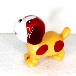 Può includere: Un cane giocattolo di plastica giallo con macchie rosse e una testa rossa e bianca. Il cane ha un'espressione giocosa.