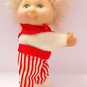 Puede incluir: Muñeca vintage con rostro redondo, ojos azules y cabello rubio claro de hilo. La muñeca viste un atuendo a rayas rojas y blancas con cuello rojo y zapatos blancos. El brazo de la muñeca está extendido.