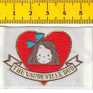 Può includere: Etichetta in tessuto bianco con un cuore ricamato in oro che presenta una ragazza con un fiocco blu. Il testo "The Vaudeville Div" è ricamato in oro sotto il cuore.