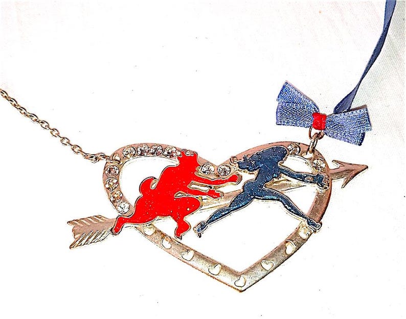 Op de afbeelding: Een gouden ketting met een hartvormige hanger met een rood en blauw emaille ontwerp van een vrouw en een hond. De hanger is bevestigd aan een zilveren ketting en heeft bovenaan een blauwe strik.