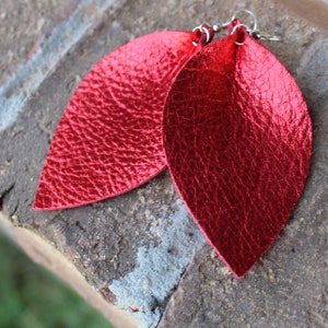 May include: A pair of red leaf-shaped earrings. The earrings have a textured, metallic finish and are suspended from silver-colored ear wires. The earrings are displayed on a textured, brick-like surface.