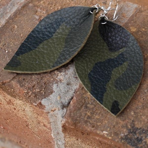 May include: A pair of leaf-shaped earrings with a camouflage pattern in shades of green and black. The earrings are made of textured material and have silver-coloured ear wires. The earrings are displayed on a brick surface.