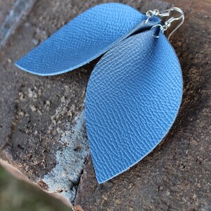 May include: A pair of blue leather leaf-shaped earrings with silver-colored hooks. The earrings have a textured surface and are displayed on a rough, dark brown surface.