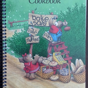 Peut inclure: Un livre de cuisine à reliure spirale intitulé Family Favourites Cookbook de la Calgary First Church of the Nazarene. La couverture présente une illustration de deux ours à une vente de pâtisseries avec des panneaux pour gâteaux, muffins et tartes.