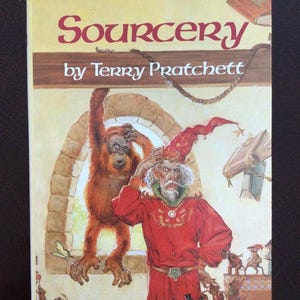 May include: Book cover for "Sourcery" by Terry Pratchett. The cover art features a wizard in a red robe, an orangutan, and various small creatures. The title is in a stylized font, with a light brown background.