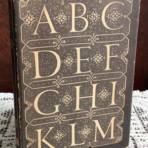 May include: A black hardcover book titled A Book of Scripts with the text A King Penguin at the bottom. The front cover features a gold-colored alphabet from A to M arranged in four rows, decorated with intricate floral and geometric patterns.