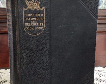 May include: A vintage cookbook titled "Household Discoveries and Mrs. Curtis's Cook Book." The cover is black with gold lettering and an illustration of a ship. The book appears to be well-worn, suggesting it is an antique.