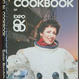 May include: The Official Cookbook of Expo 86 by Susan Mendelson is a spiral-bound book featuring a cover image of a person wearing a white space suit with dark circular fasteners. The title text is printed in white against a dark background.