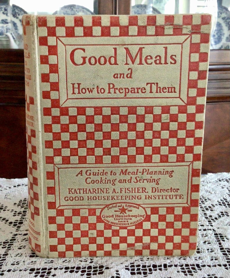 May include: A vintage cookbook titled "Good Meals and How to Prepare Them" with a red and white checkered cover. The book is a guide to meal planning, cooking, and serving by Katharine A. Fisher, Director of the Good Housekeeping Institute. The book has a "Tested and Approved" Good Housekeeping Institute seal.