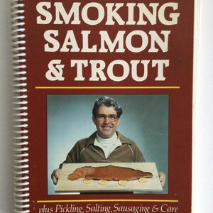 May include: A cookbook titled "SMOKING SALMON & TROUT" with a brown cover and white spiral binding. The cover features a photo of a man holding a wooden board with smoked fish and lemon slices. Additional text includes "plus Pickling, Salting, Sausaging & Care" and the author's name.