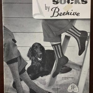May include: A vintage advertisement for socks by Beehive featuring a black dog and two pairs of legs wearing patterned socks. One pair displays a chevron design, while the other features vertical stripes. The text reads men's socks by Beehive.