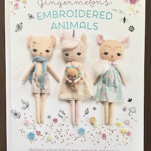 May include: The cover of a book, "Gingermelon's Embroidered Animals", displays three fabric animal dolls: a bear, a unicorn, and a deer. The dolls are light-toned with embroidered features and dressed in fabric clothing. The book's subtitle reads "Heirloom animal dolls to sew, embellish and treasure."