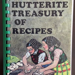 Peut inclure: Un livre de cuisine à reliure spirale intitulé The Hutterite Treasury of Recipes avec une spirale en plastique vert. La couverture présente un dessin de deux personnes portant des foulards à pois pétrissant de la pâte sur une table.