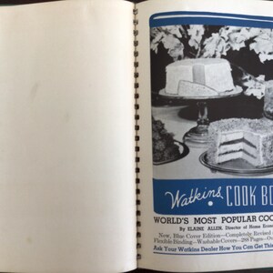 Puede incluir: Un libro de cocina Watkins de encuadernaci&oacute;n en espiral abierto con una cubierta azul y blanca que presenta fotograf&iacute;as en blanco y negro de pasteles en capas sobre pedestales. El texto dice Watkins COOK BOOK, WORLD'S MOST POPULAR COOK BOOK.