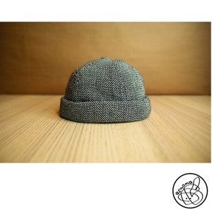 May include: A gray and white speckled beanie hat with a folded brim. The hat is made of a textured fabric and sits on a light-colored wooden surface. The background is a solid, warm brown.