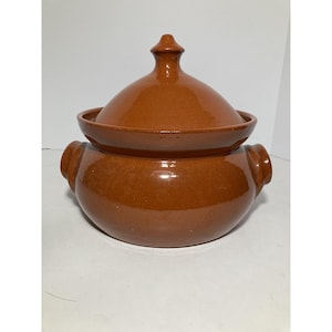 Puede incluir: Una olla de cerámica marrón con tapa y dos asas laterales. La olla tiene una base redondeada y una tapa con un pequeño pomo puntiagudo. La olla está esmaltada y tiene un acabado brillante. Probablemente se utiliza para cocinar o servir alimentos.