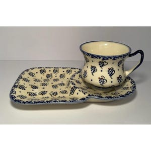 May include: A cream-colored ceramic teacup and saucer set with a matching serving tray. The set features a blue grape pattern and a blue trim. The teacup has a curved handle. The tray is rectangular with a curved section for the cup.