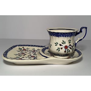 May include: A ceramic teacup and saucer set with a matching rectangular plate. The set features a cream-colored base with blue floral patterns and trim. The cup has a curved handle and the set is designed for serving tea.