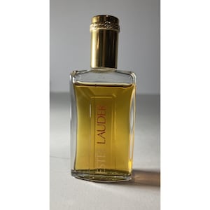 May include: A rectangular glass perfume bottle with a gold-coloured cap and a gold-coloured band around the neck. The bottle contains a golden-yellow liquid and has the words "ESTEE LAUDER" printed vertically in red.
