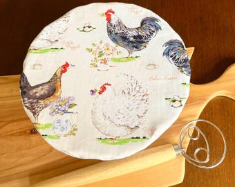 May include: A round, light-colored fabric bread cover with a rooster and hen pattern. The cover features illustrations of chickens, eggs, and floral arrangements. A wooden handled dough whisk is in the foreground, resting on a wooden cutting board.