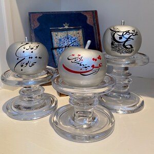 May include: Three silver apples with black calligraphy on a white background are displayed on clear glass candle holders. The apples are on a white surface.