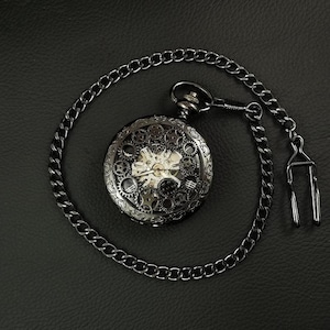 Puede incluir: Un reloj de bolsillo plateado con una cadena negra y engranajes intrincados visibles a través de la esfera.