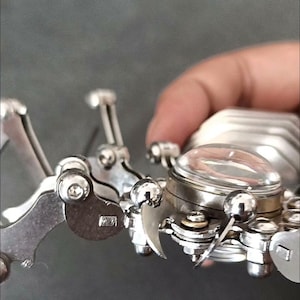 May include: A silver metal robotic spider with a clear glass dome on its back. The spider has multiple articulated legs and a detailed design.
