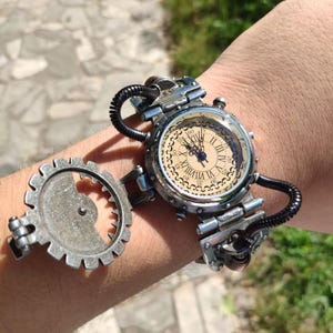 Op de afbeelding: Een zilveren steampunk-stijl polshorloge met een ronde, cr&egrave;mekleurige wijzerplaat en Romeinse cijfers voor de uren. Het horloge heeft een groot, zilveren tandwiel dat aan de band is bevestigd.