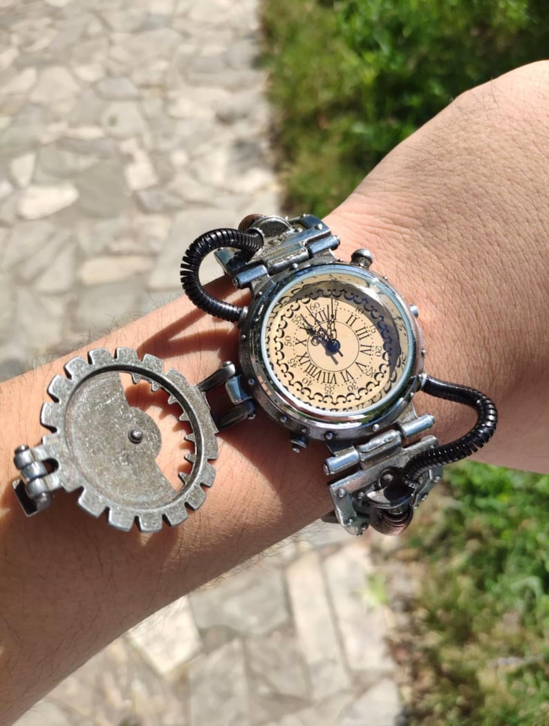 Op de afbeelding: Een zilveren steampunk-stijl polshorloge met een ronde, cr&egrave;mekleurige wijzerplaat en Romeinse cijfers voor de uren. Het horloge heeft een groot, zilveren tandwiel dat aan de band is bevestigd.