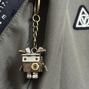 Peut inclure: Un porte-clés robot de style steampunk en métal et en bois. Le robot a un corps carré, des accents en laiton, et est attaché à une chaîne et un porte-clés en laiton. Le design du robot comprend des ressorts, des boulons et d'autres détails mécaniques.