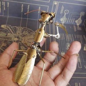 May include: A gold-toned metal praying mantis sculpture with intricate details and movable parts. The sculpture is made of brass and has a steampunk aesthetic.