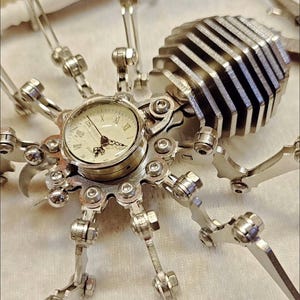 May include: A silver metal spider clock with a round clock face and Roman numeral markings. The spider has eight legs and a segmented body.