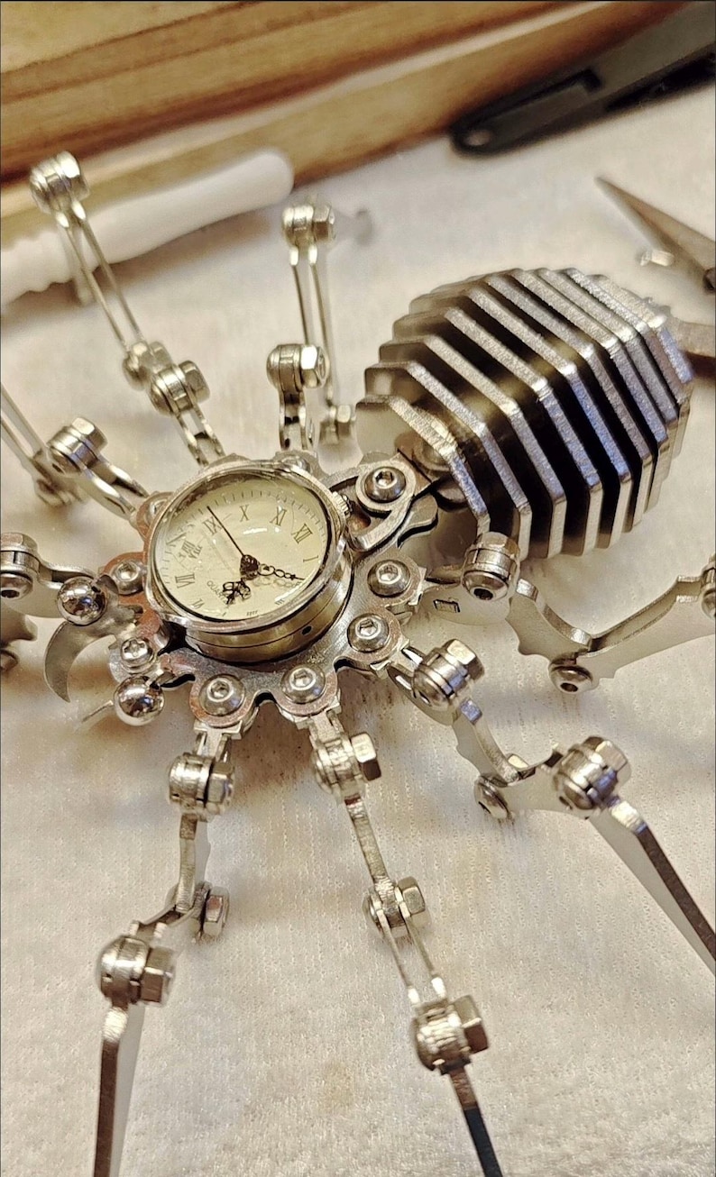 May include: A silver metal spider clock with a round clock face and Roman numeral markings. The spider has eight legs and a segmented body.