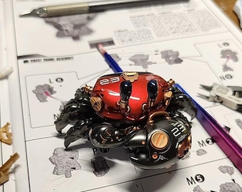 Può includere: Un kit modello a forma di granchio rosso e nero con il numero "23" sul guscio, circondato da strumenti e istruzioni di montaggio. Il modello ha dettagli dorati e neri.